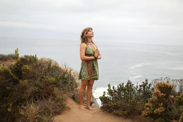 A barefoot woman in a dress presses her hands together in prayer, eyes closed and tilted to the sky, while on a beach.