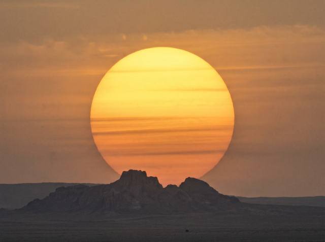 Large golden sun setting behind the dark silhouette of desert rock formations under an orange sky.