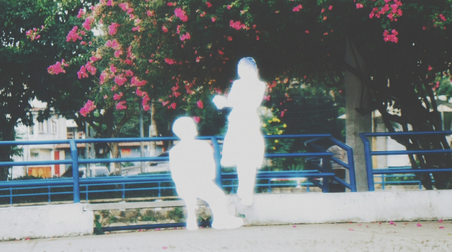 Two heavily overexposed white silhouettes of a child and an adult standing near a flowering tree with pink blossoms and blue railings in a park.