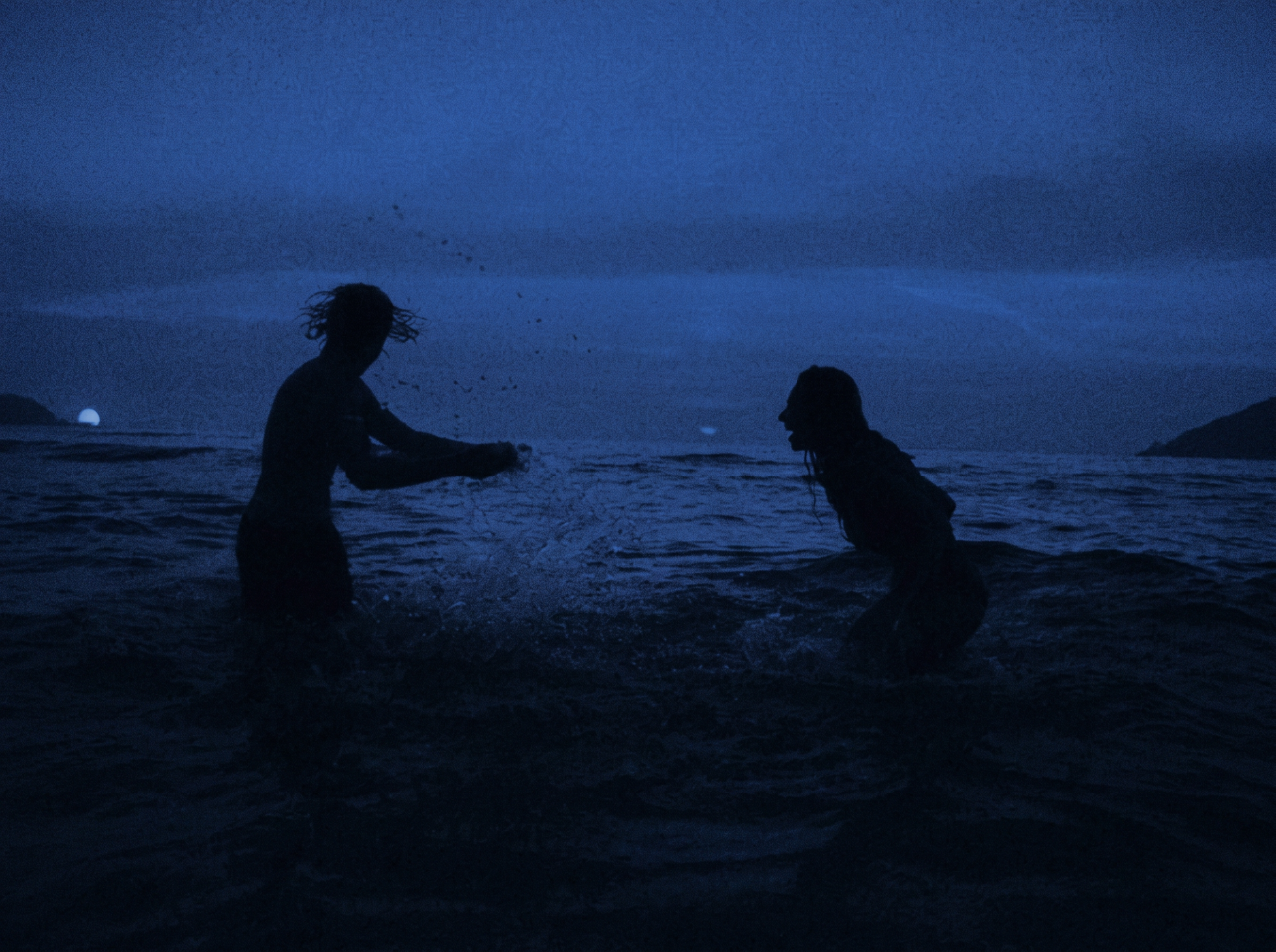 Silhouettes of two people splashing each other in the ocean at dusk, blue twilight sky and distant hills in the background.