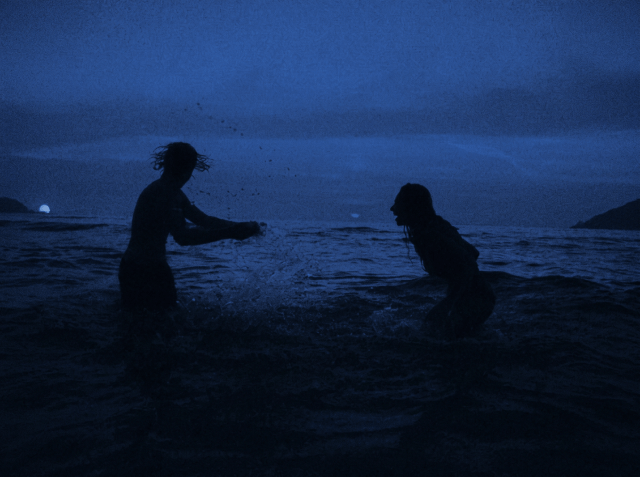Silhouettes of two people splashing each other in the ocean at dusk, blue twilight sky and distant hills in the background.
