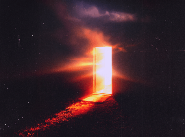 A luminous open door radiating intense orange and white light stands alone in a dark landscape under a dramatic cloudy night sky.
