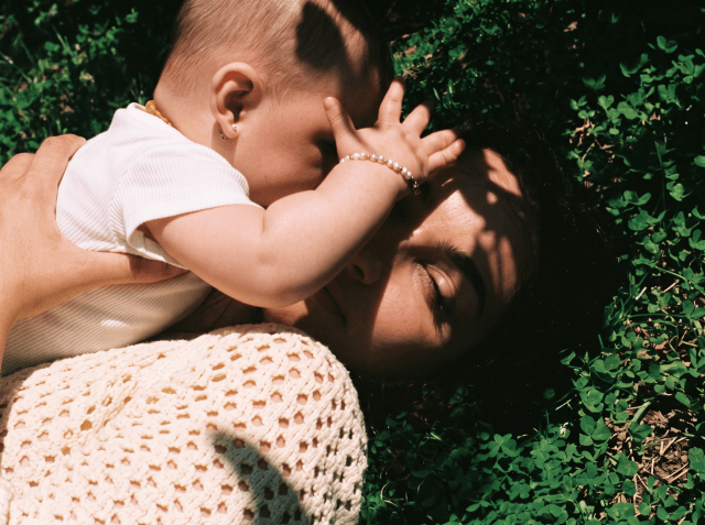 Mother lying in a clover field holding her baby, who reaches a small hand toward her face.