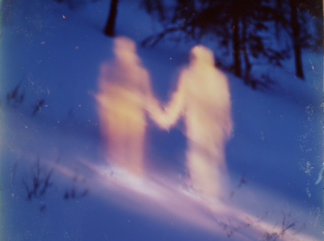 Blurry, luminous silhouettes of two people holding hands in a snowy blue-toned scene, shot in a dreamy out-of-focus Polaroid style.