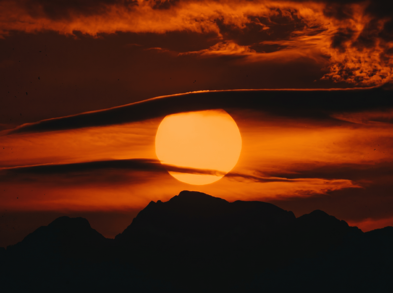 Large orange sun setting behind a dark mountain silhouette against a deep red and orange cloudy sky.