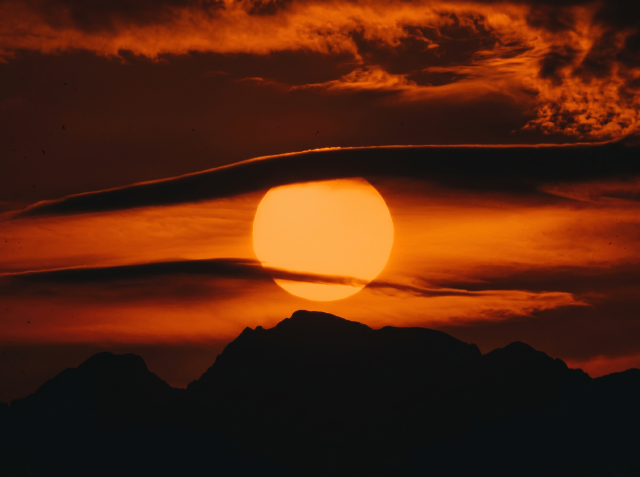 Large orange sun setting behind a dark mountain silhouette against a deep red and orange cloudy sky.