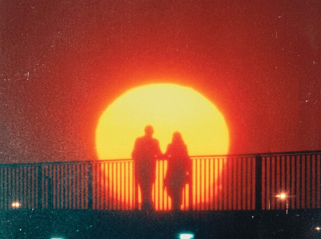 Silhouettes of two people holding hands on a bridge railing against a large glowing sun and deep red sky.