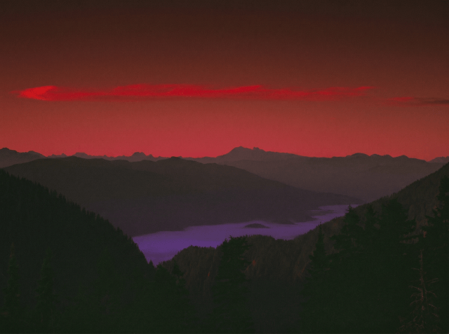 Deep red and crimson sunset sky over silhouetted mountain ridges with a purple-hued lake nestled in a forested valley below.
