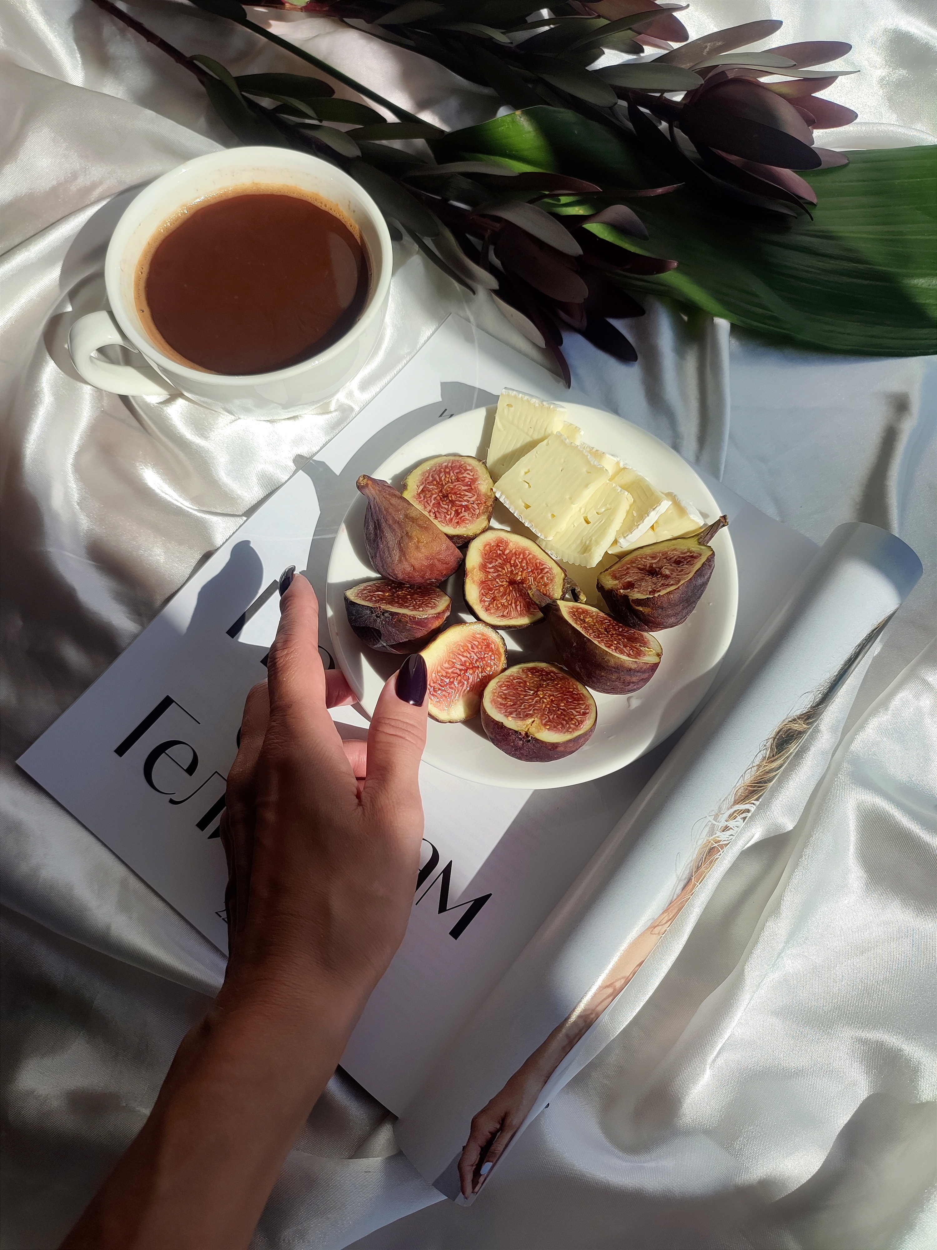 Overhead view of a hand holding a plate of sliced figs and cheese next to a cup of black coffee on white silk fabric with dark flowers nearby.